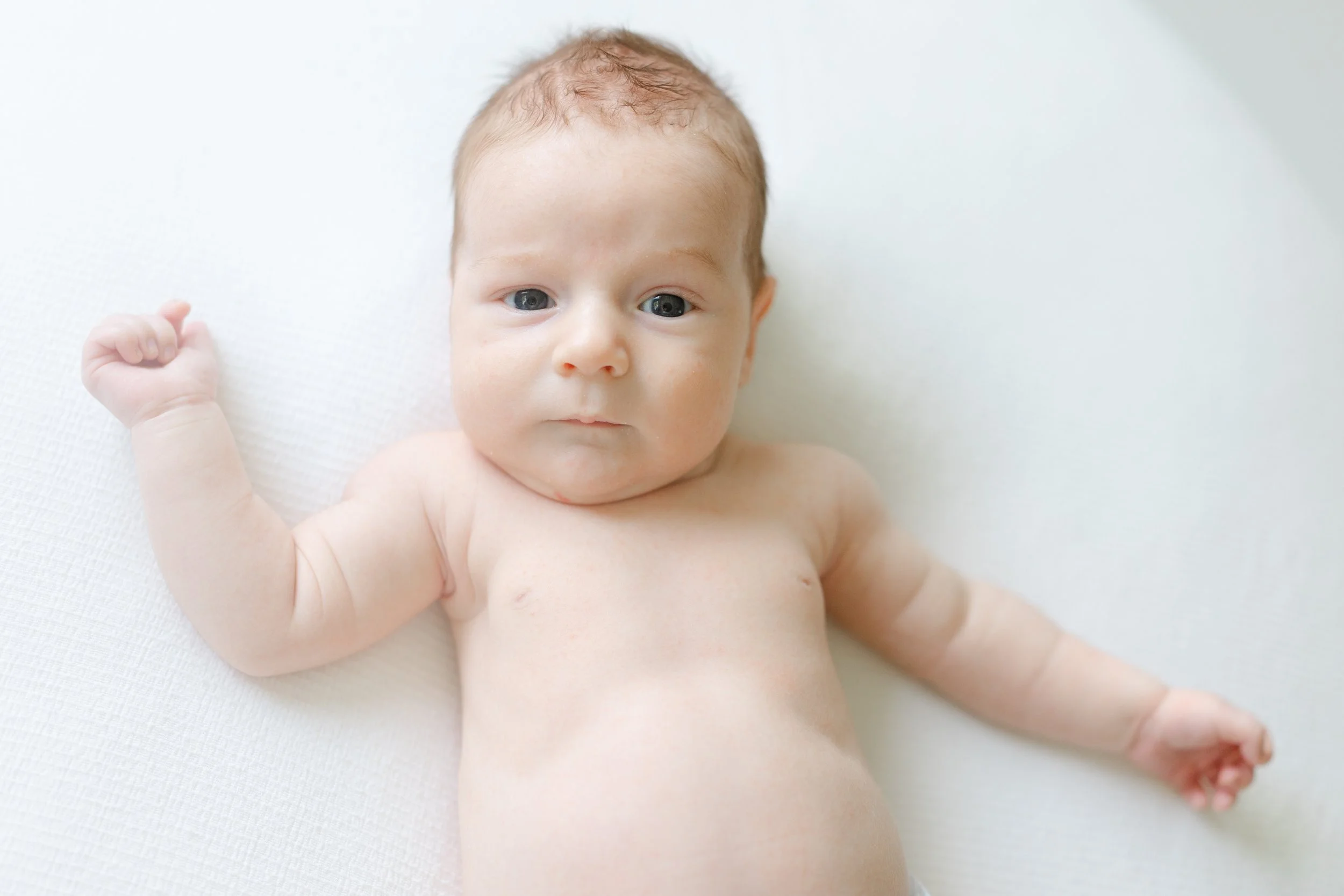 Close-up of a naked baby with light skin and blue eyes lying on a white textured surface, looking into the camera, with one arm raised and curled fist.