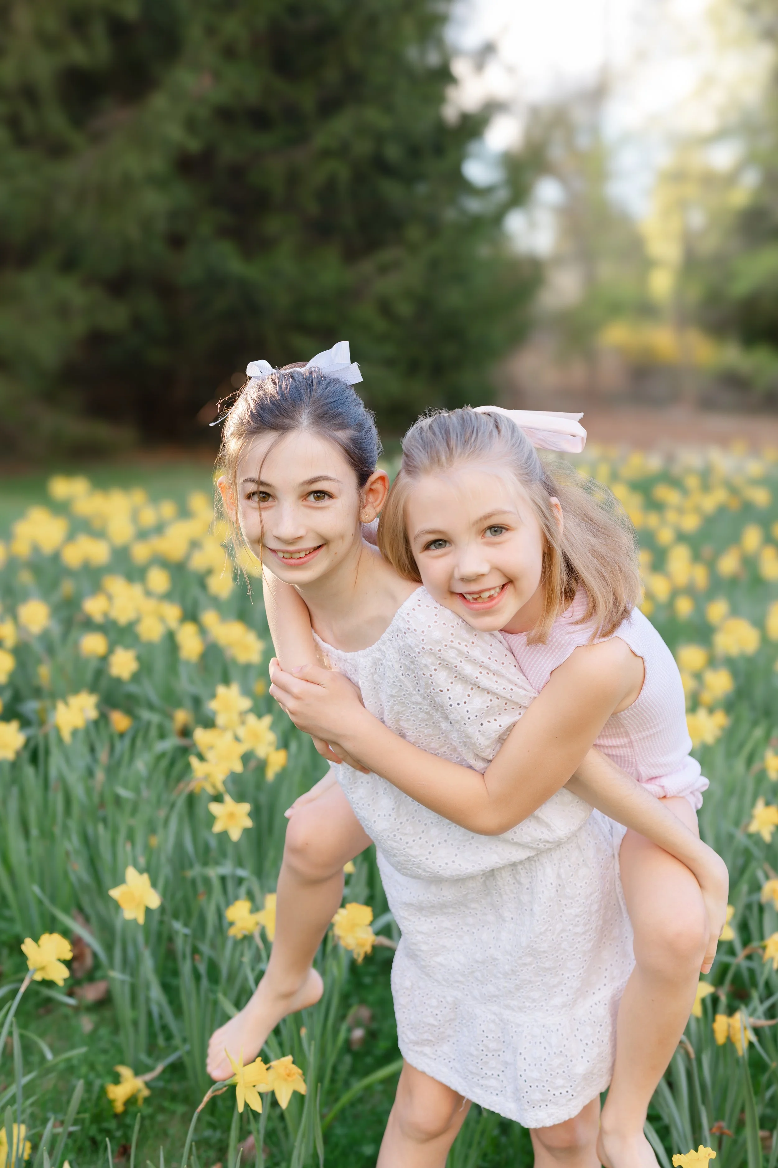 Two young girls with light skin and long hair, one with dark hair and one with blond hair, smiling and embracing each other in a field of yellow flowers with a blurred green trees background.