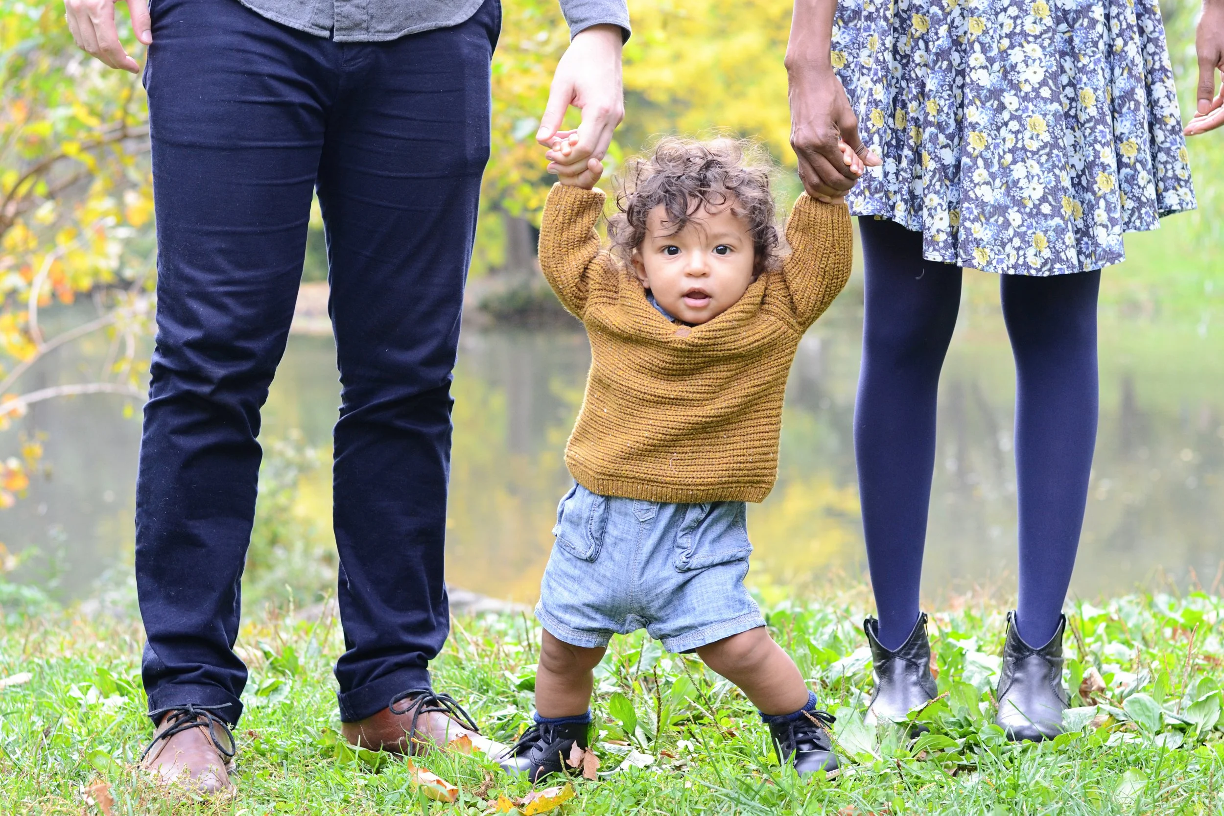 A young child with curly hair holding hands with two adults, one on each side, in a park with green grass and trees in the background. The child is wearing a brown sweater and gray shorts, while the adults are dressed in dark pants and floral dresses