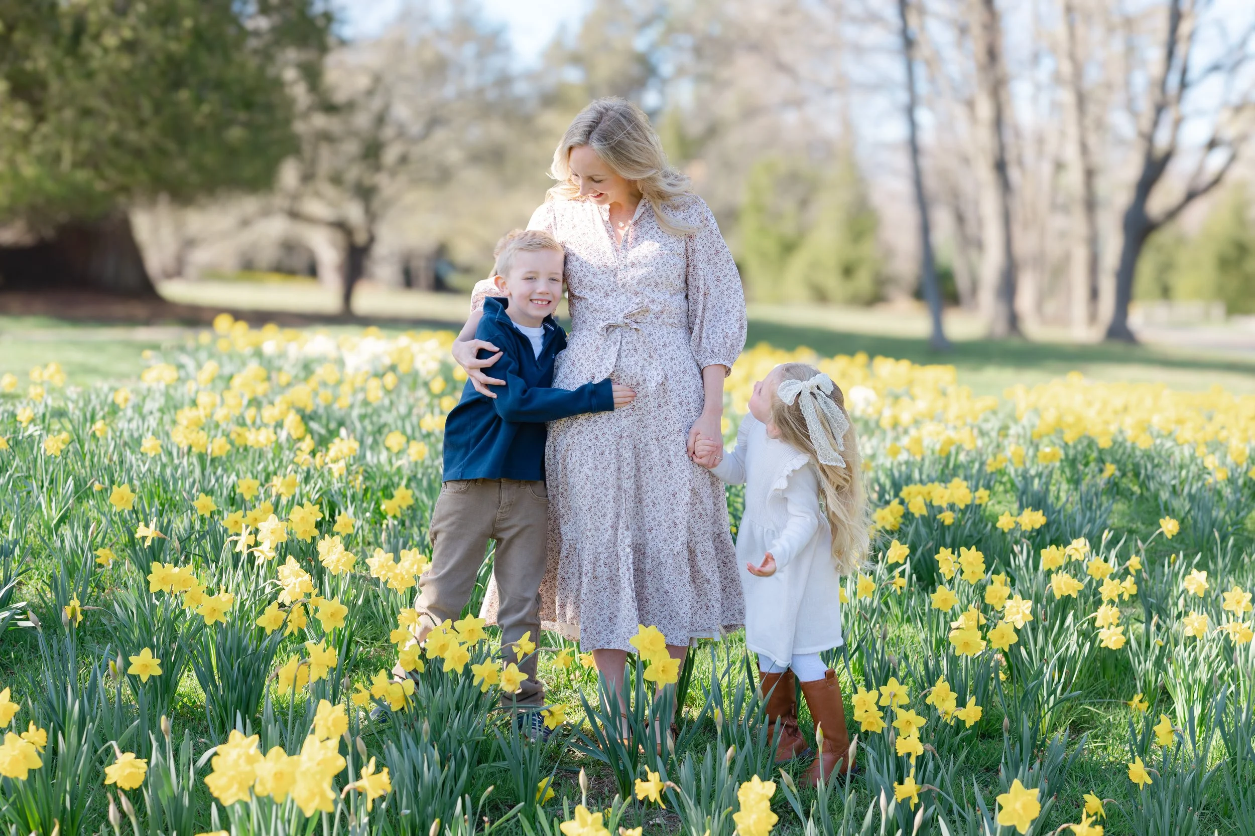 A woman with blonde hair in a floral dress standing in a field of yellow flowers, holding hands with two children, a boy and a girl, in a park with trees in the background.