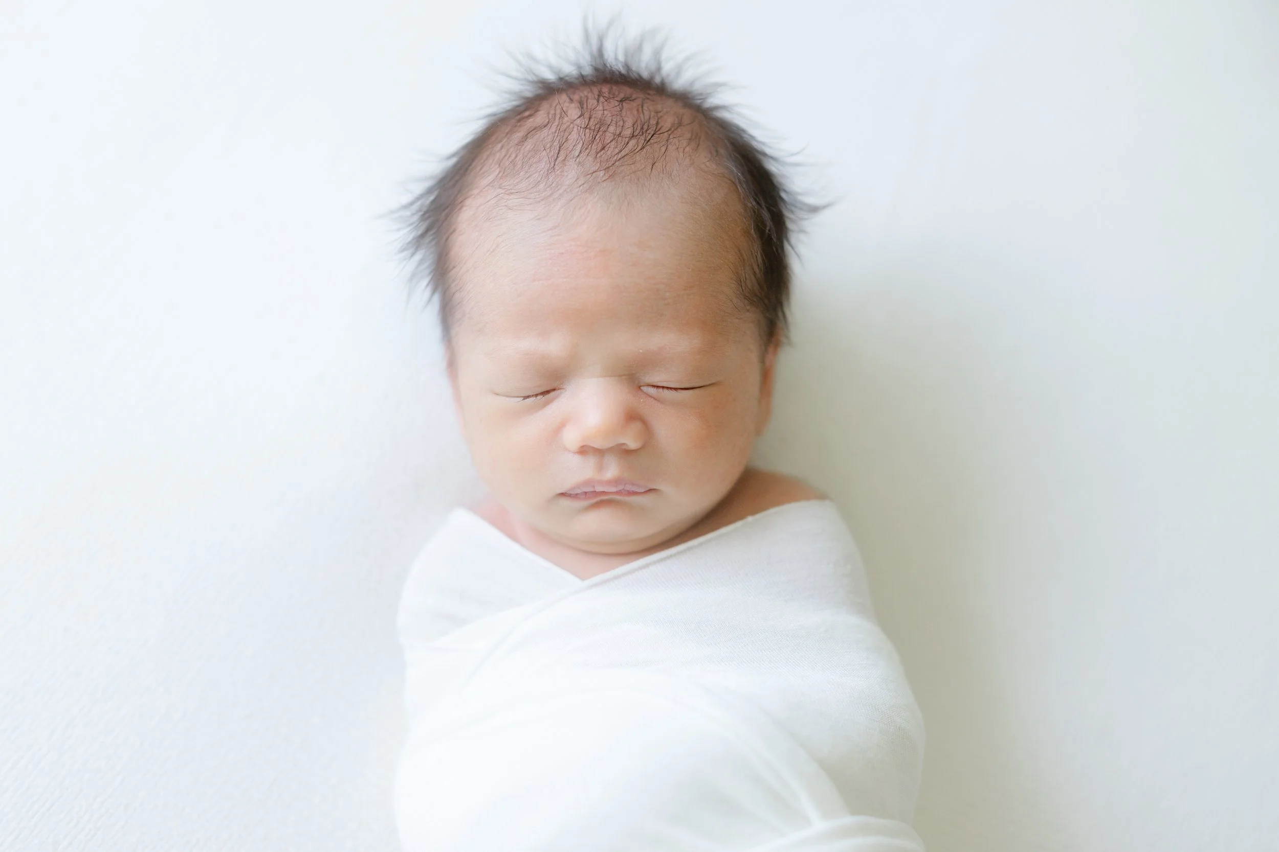 A baby with light skin and dark, sparse hair lying with eyes closed on a white surface, wearing a white shirt.