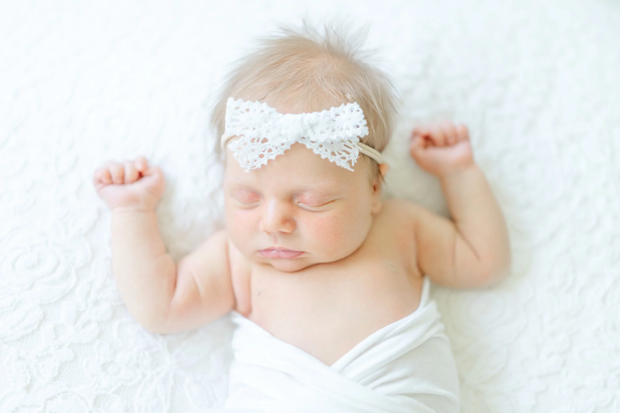 Close-up of a sleeping newborn baby with a lace headband, lying on a white textured blanket.