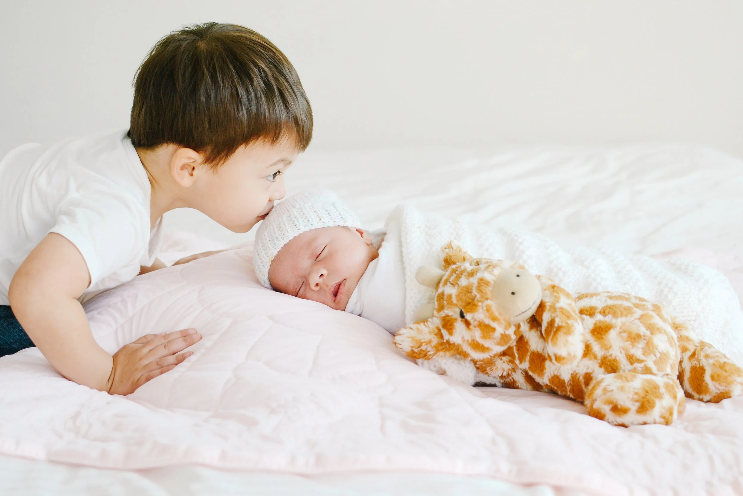 A young boy with brown hair leaning over and kissing a sleeping newborn baby who is dressed in a white knit hat and wrapped in a white blanket, lying on a bed with a stuffed giraffe toy nearby.