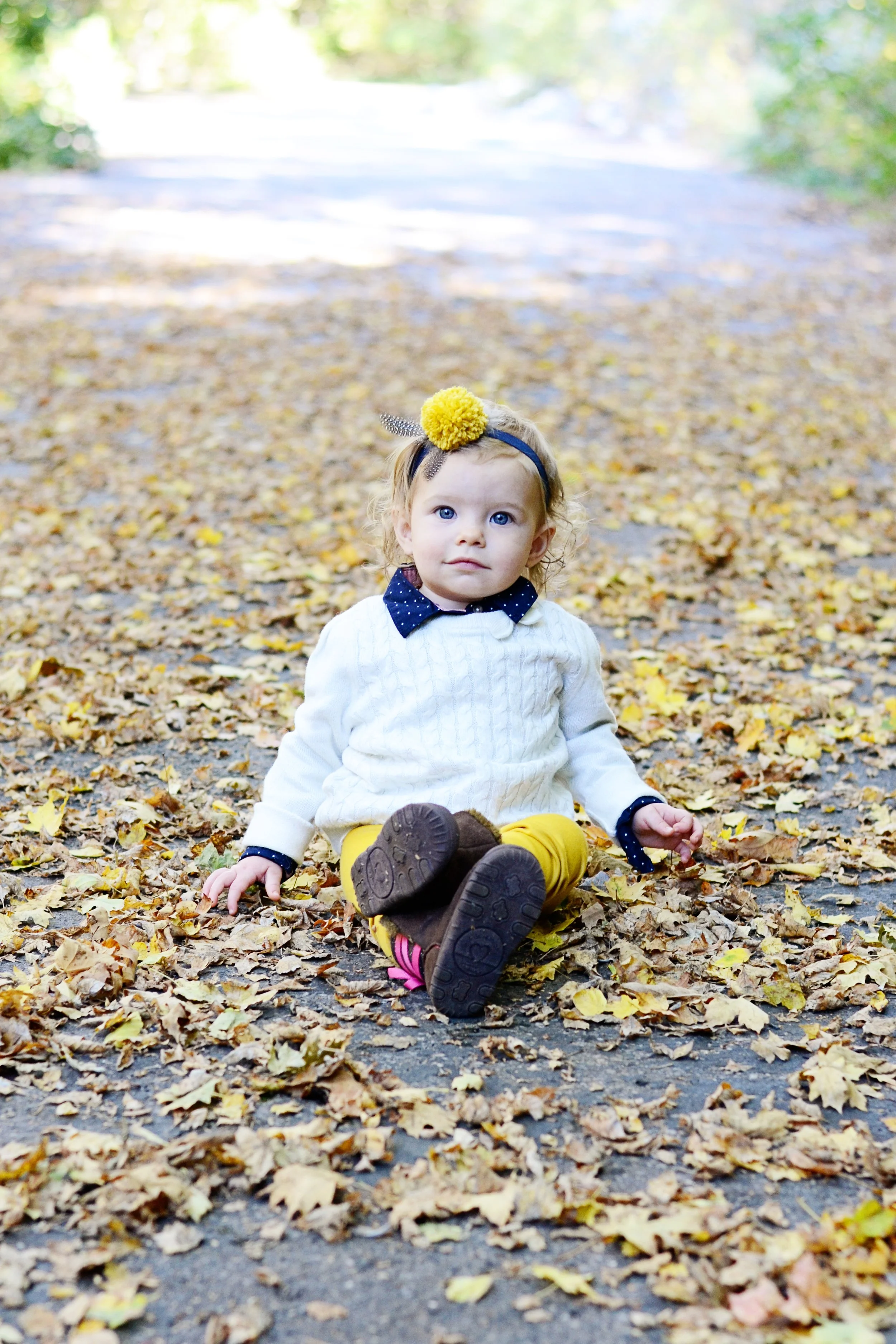 Young girl with blonde hair sitting on the ground covered in yellow leaves during autumn, wearing a cream sweater, navy polka-dotted shirt, yellow pants, brown shoes, and a black headband with a yellow pom-pom and black and white feather. She's outdo