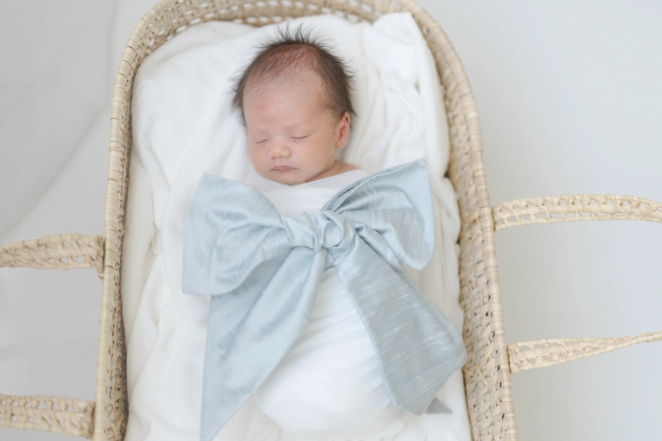 A sleeping newborn baby with dark, thin hair in a basket with a white blanket and a silver satin cloth.
