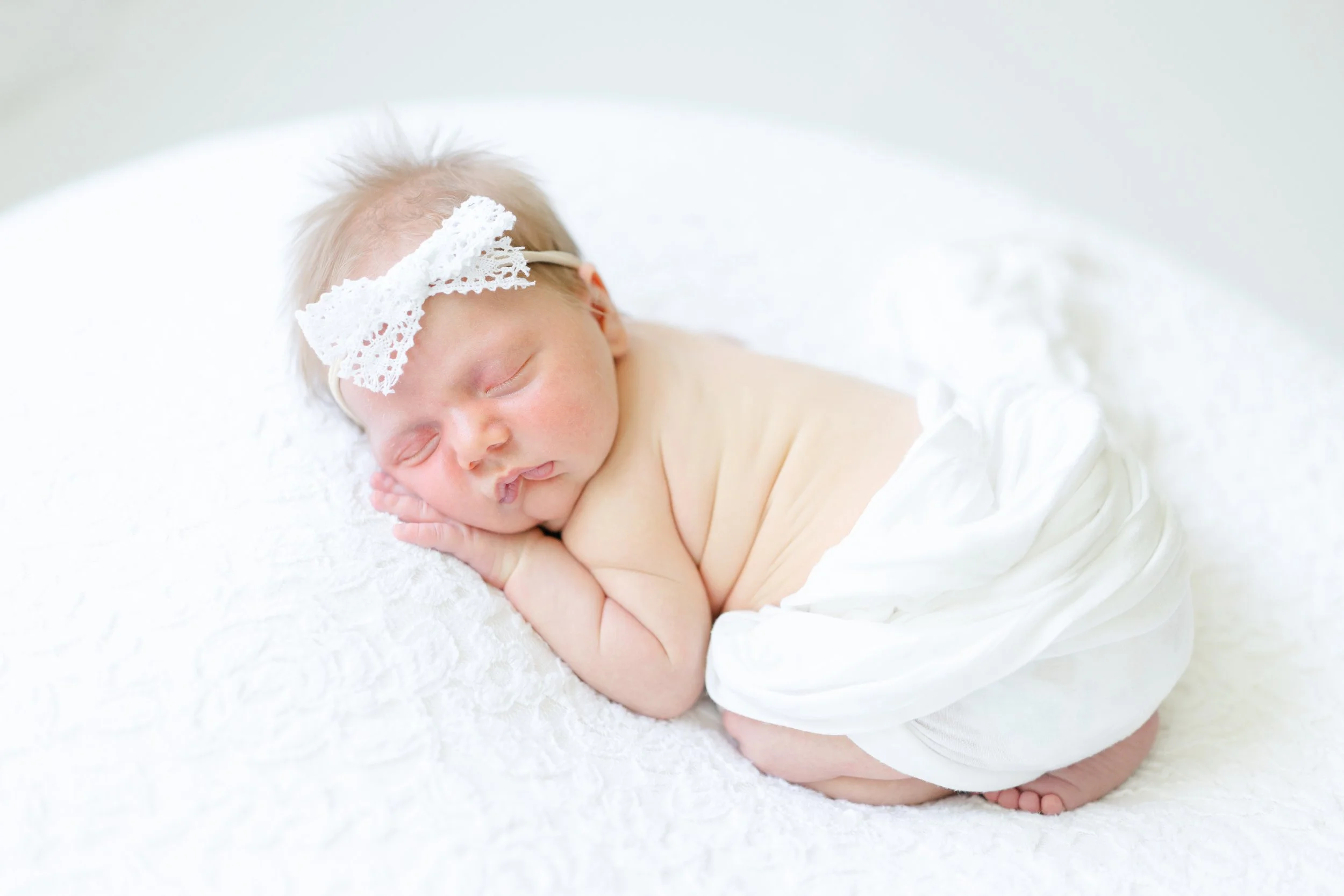Young baby with blonde hair lying on a white blanket, wearing a white lace headband with a bow, sleeping peacefully with hands under petal.