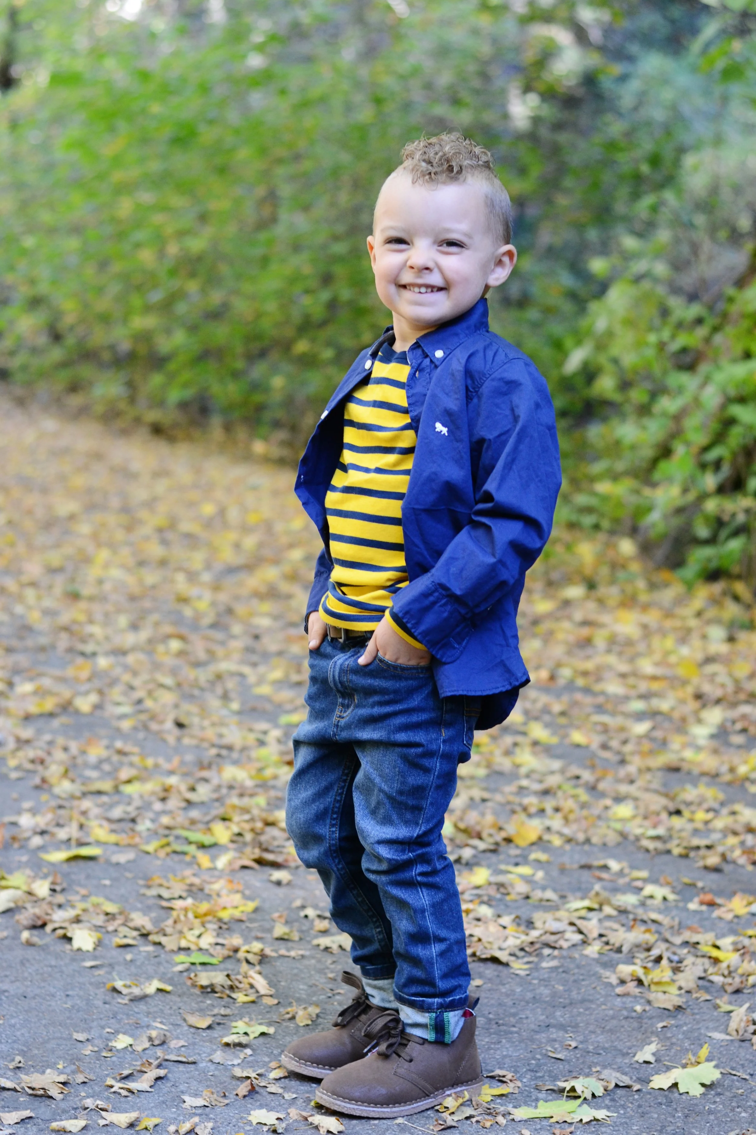 A young boy wearing a blue jacket, yellow and navy striped shirt, jeans, and brown boots, standing outdoors on a leaf-covered path with a background of trees and green foliage.