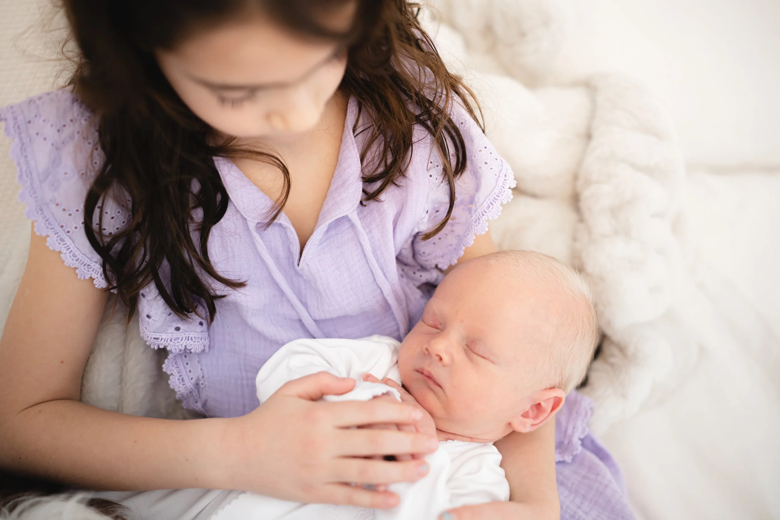 A woman with shoulder-length brown hair, wearing a lavender blouse with lace details, holding a sleeping baby with curly hair dressed in white. They are sitting on a comfortable surface with soft, white blankets.