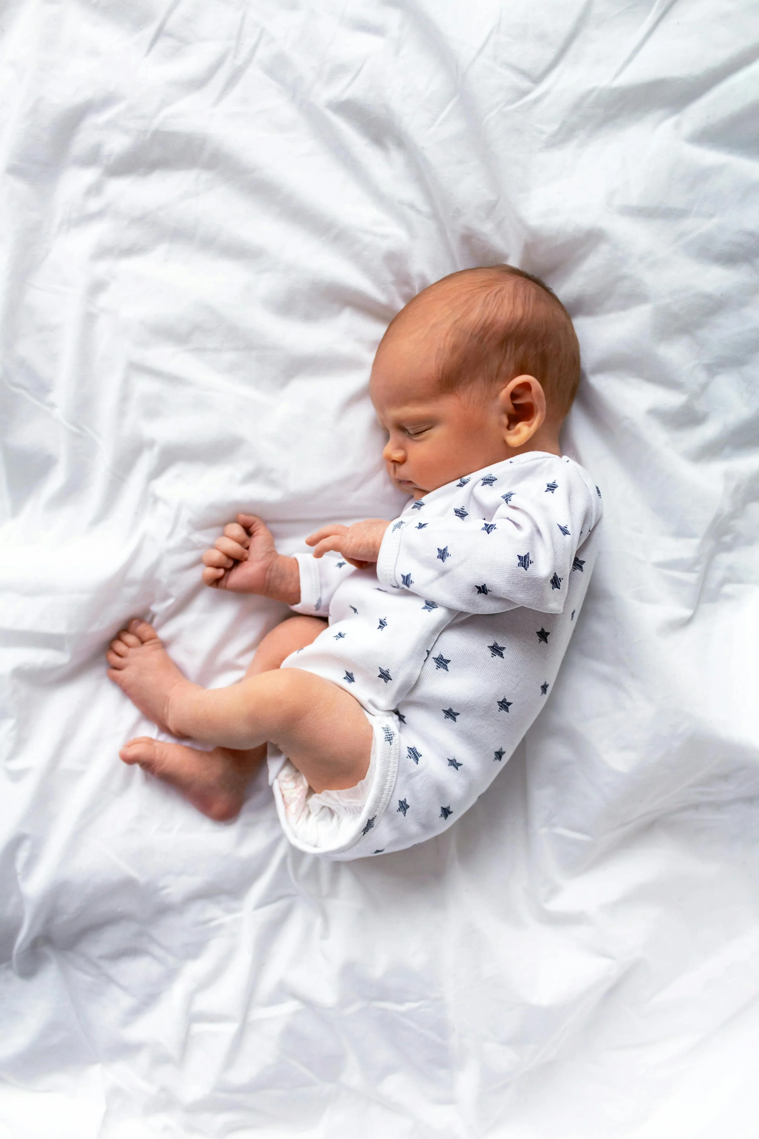 A sleeping infant with light skin and short light brown hair, lying on a white sheet, wearing a white outfit with blue star patterns.
