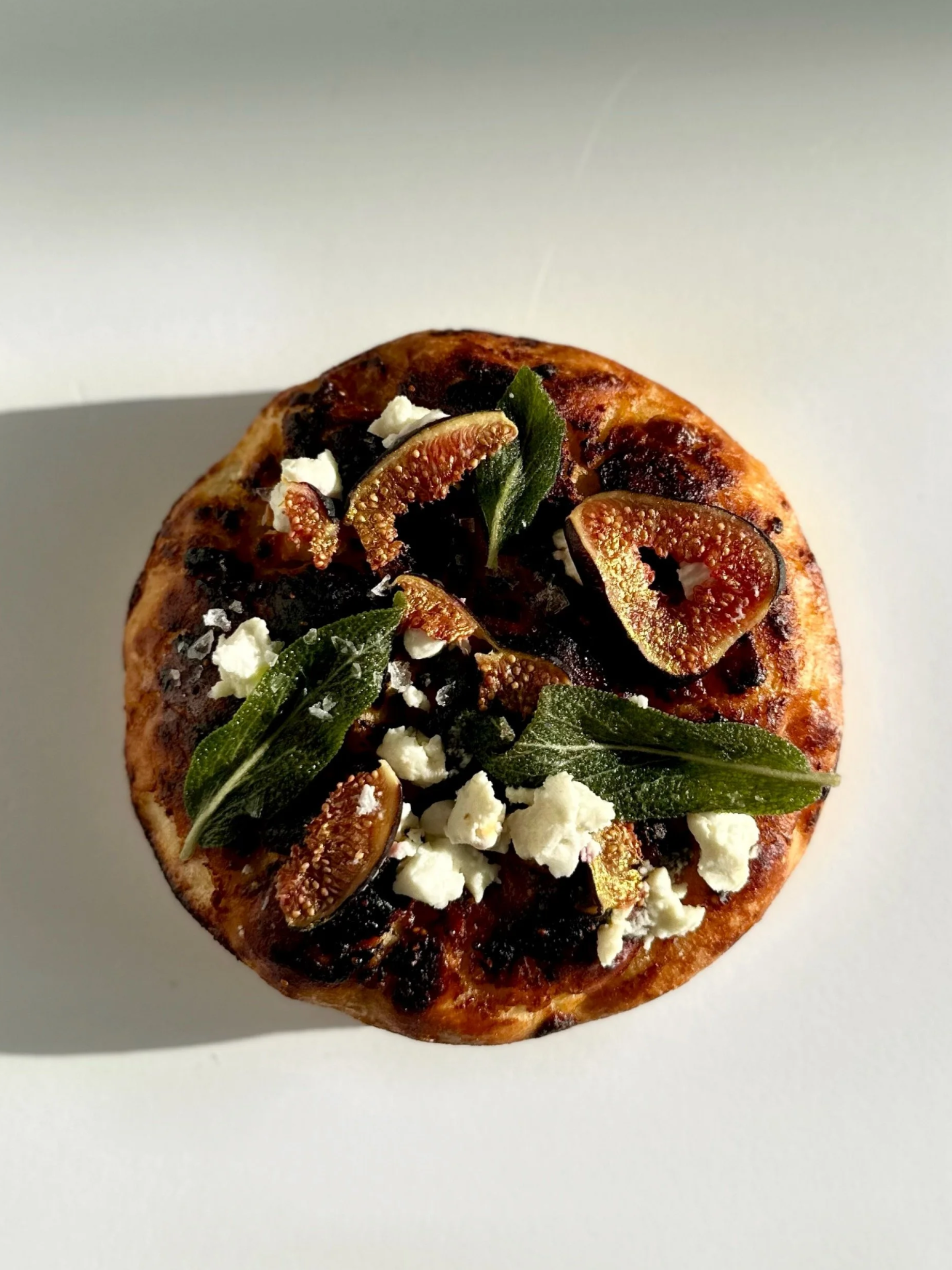 Close-up of a pizza with figs, green leaves, and white crumbles on a white surface.