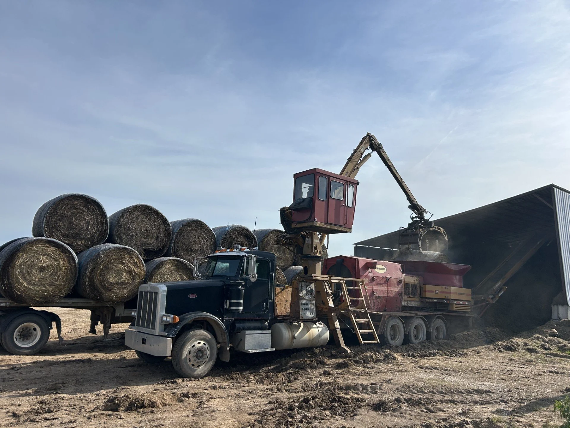 Hay bales loaded onto a truck with a mechanical loader in a farm or rural area under a partly cloudy sky.