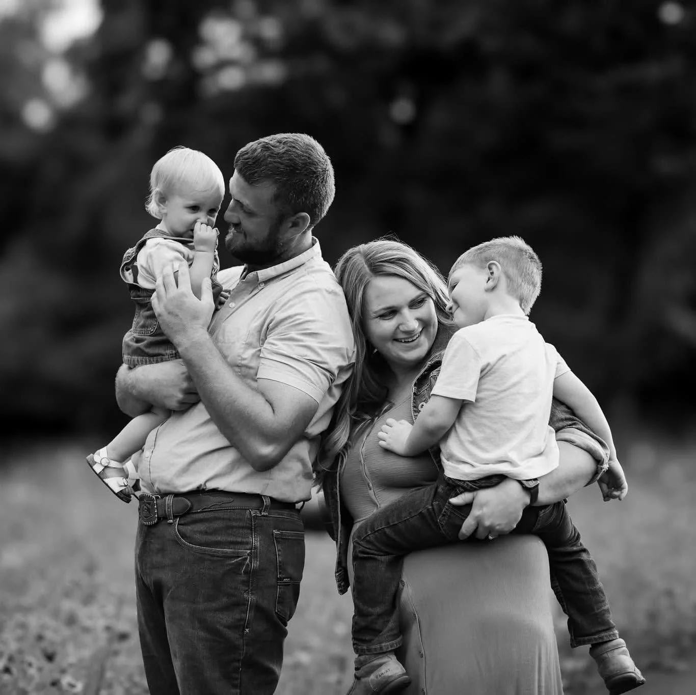 A black and white photo of a family outdoors, comprising a man holding a young girl, a woman with a smiling face holding a young boy on a stool. The family appears joyful and affectionate.