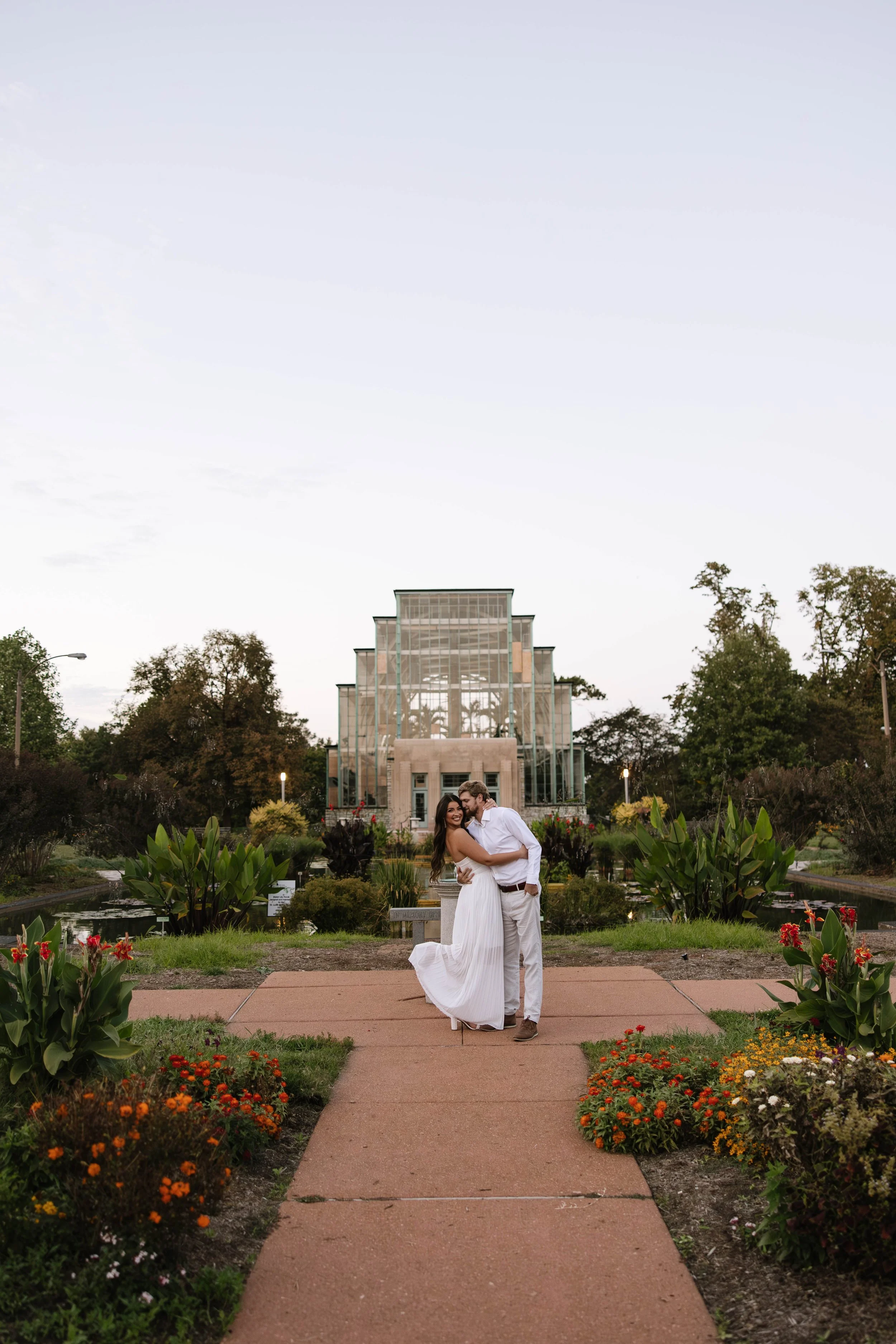 A couple in white attire embracing in front of a glass building with plants and flowers around them.