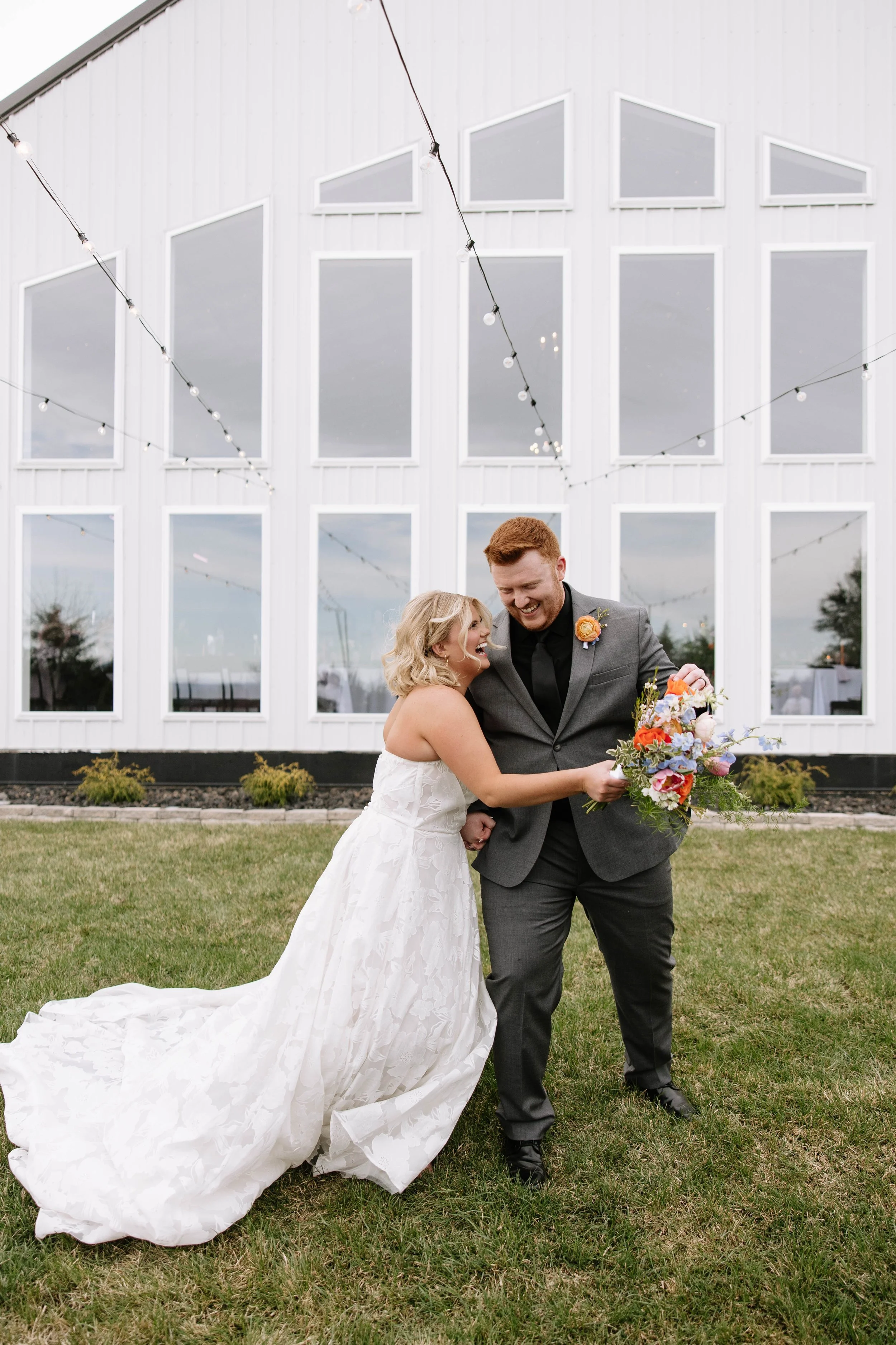 A bride and groom share a joyful moment outdoors, holding a bouquet, in front of a modern white building with large windows and string lights.