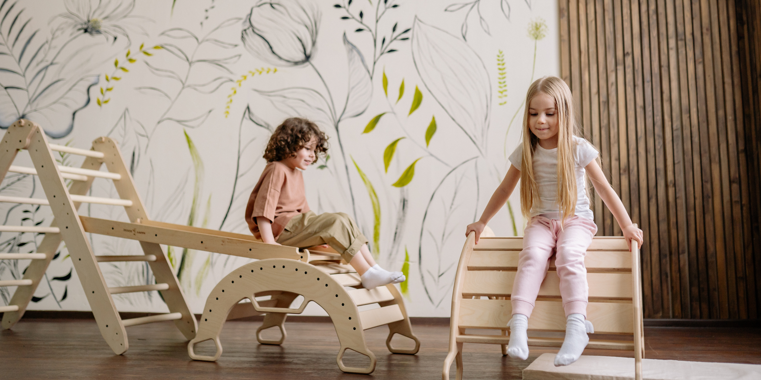 Two children playing on wooden indoor play equipment in a room with a leafy mural wall and wooden paneling.
