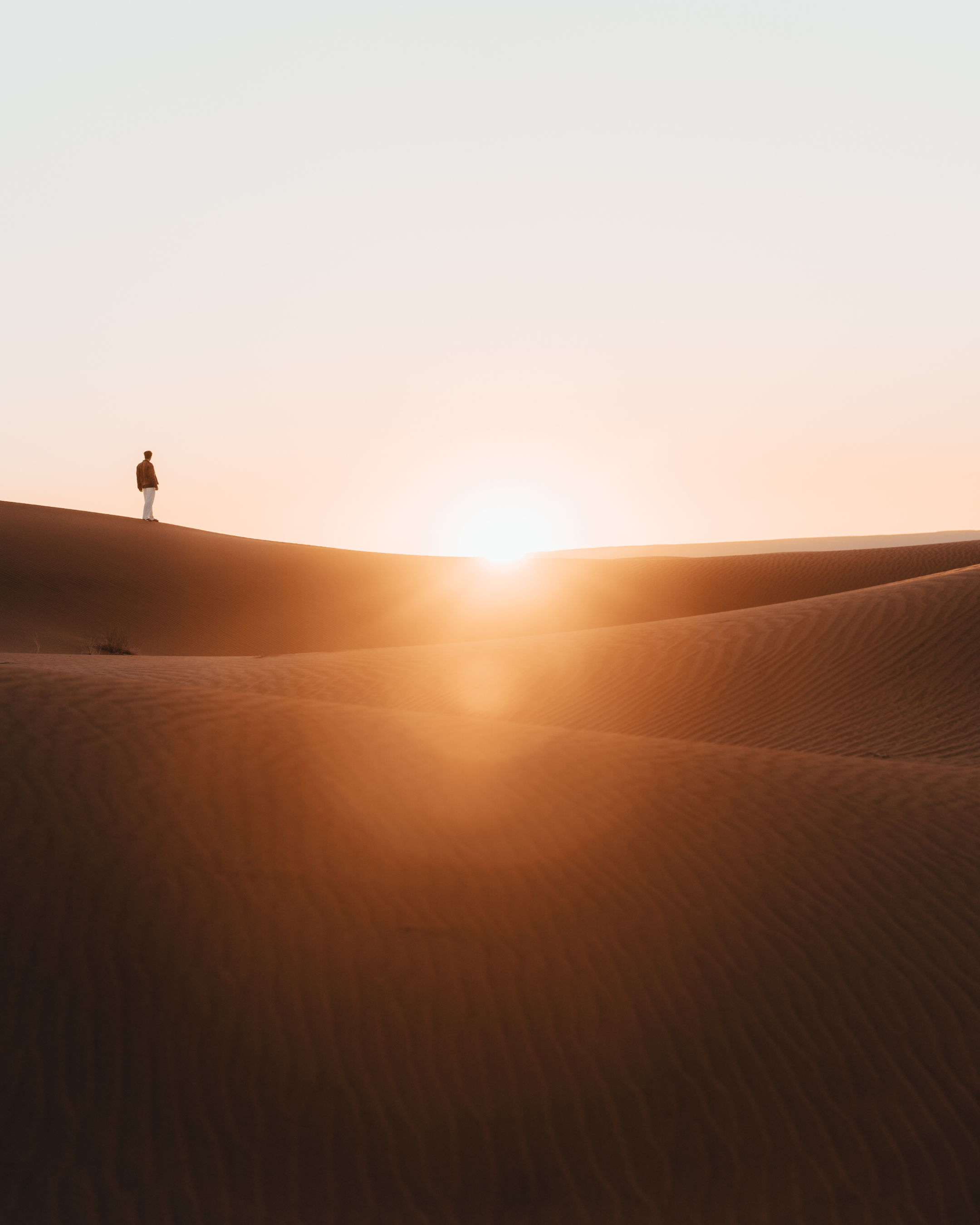 A person standing alone on a sand dune at sunset or sunrise in a desert.