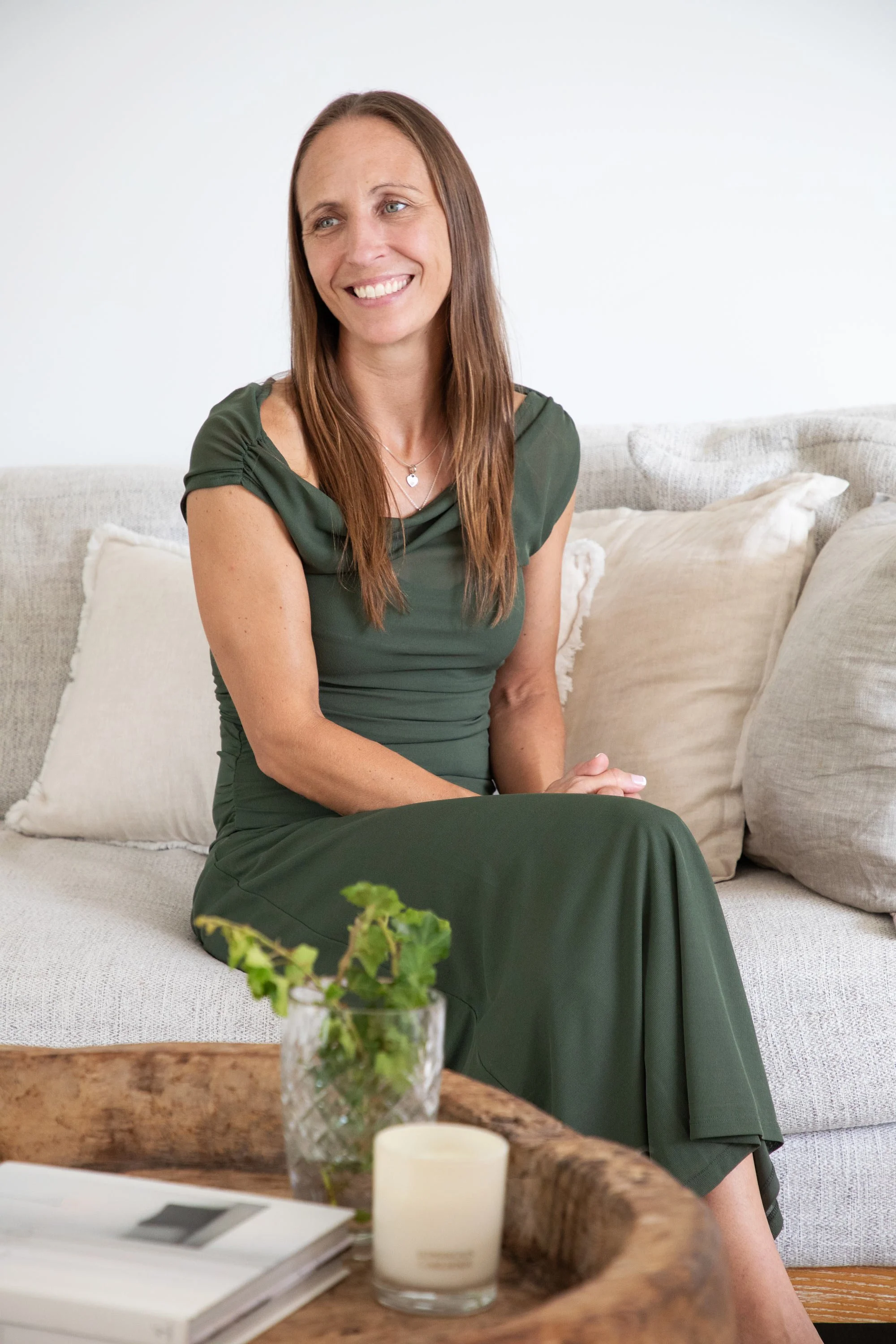 A woman with long brown hair, wearing a green dress, sitting on a beige couch with beige cushions, smiling and looking to her right. A wooden table with a book, a candle, and a vase with green foliage is in the foreground.