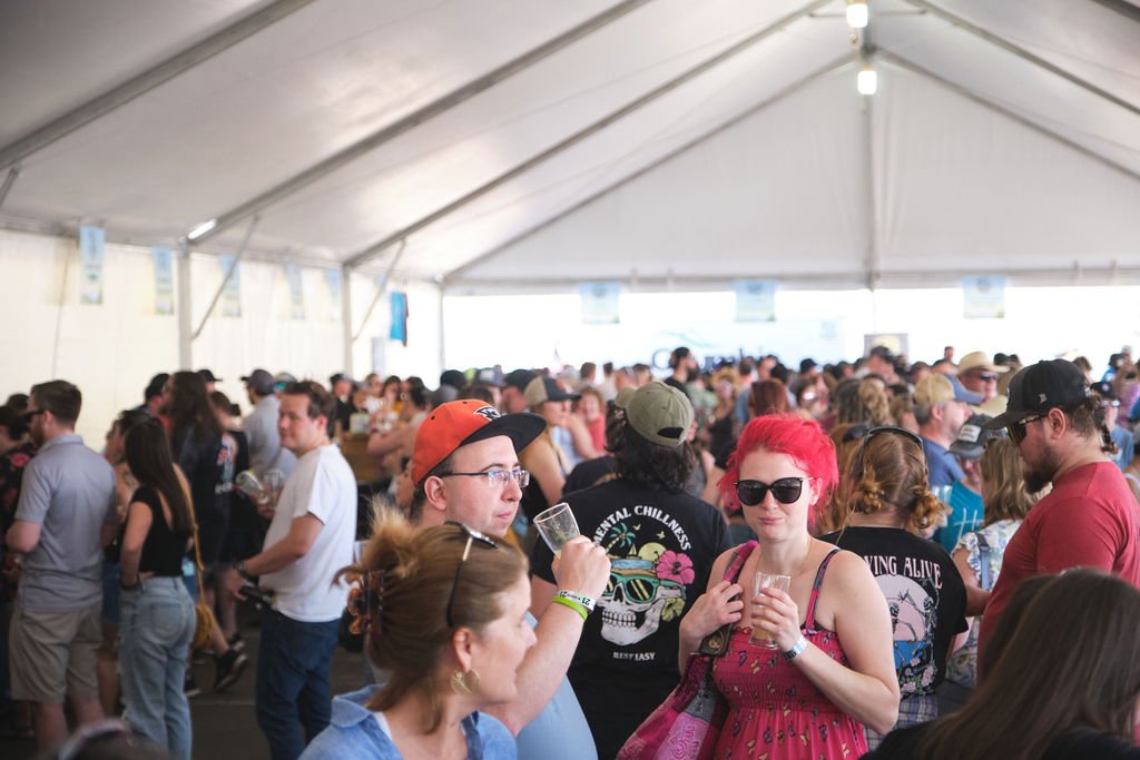 Crowd of people inside a large white event tent, many holding drinks, with some wearing sunglasses and casual summer clothing.