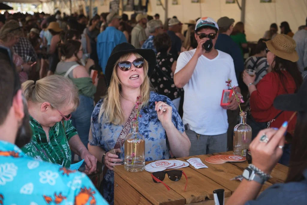 A crowd of people at an outdoor event, with a woman in the foreground wearing sunglasses, a black hat, and blue patterned shirt, holding a drink and talking. Others are wearing hats and casual clothing, some drinking and socializing.