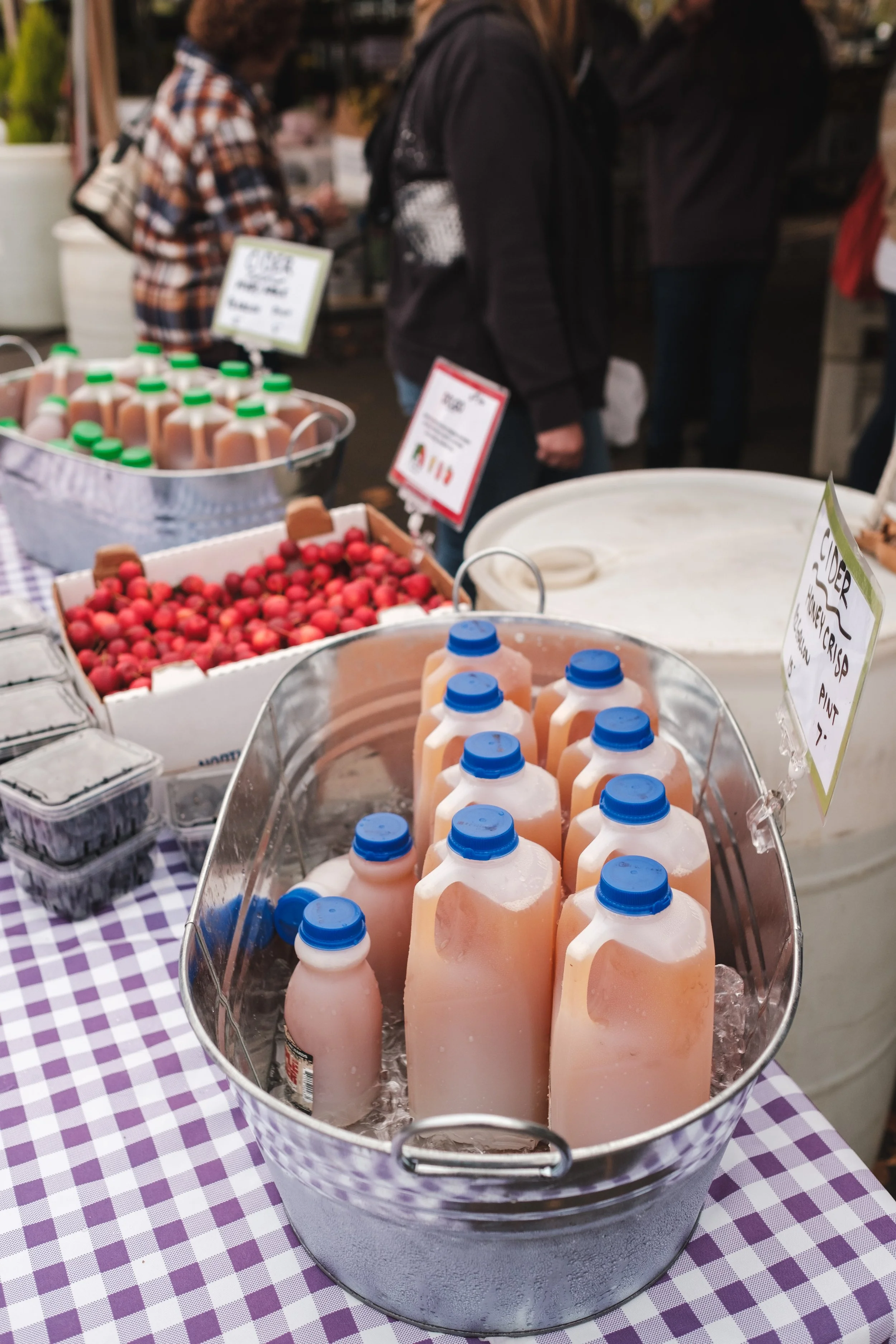 Bottles of pink lemonade in a metal bucket on a checkered tablecloth at a farmer's market.