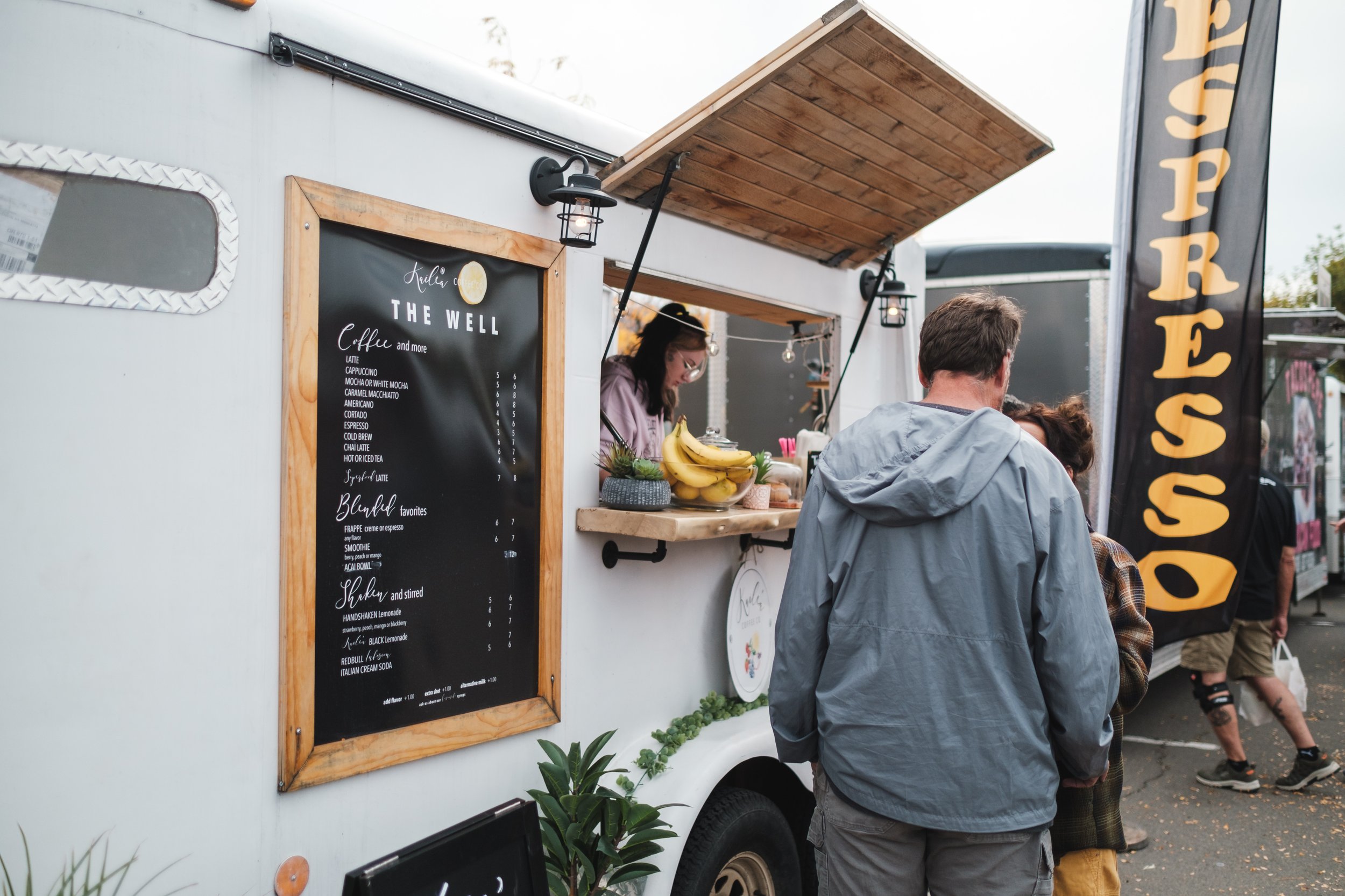 A food truck at an outdoor event with a menu board, serving coffee and drinks, and people ordering and waiting in line.
