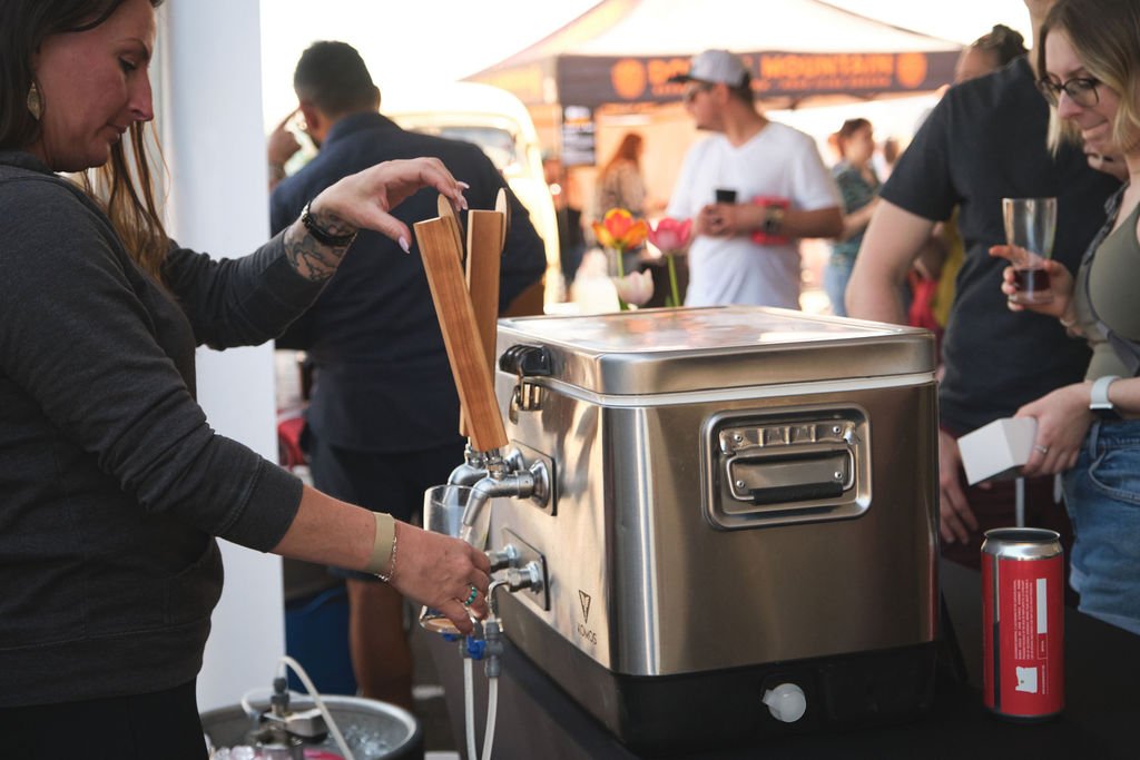 A woman serving beer from a stainless steel kegerator at an outdoor event, with people in the background enjoying food and drinks.
