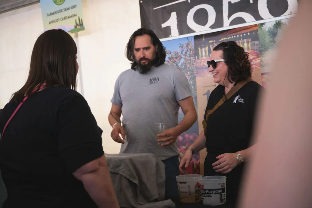 Three people engaged in conversation at a booth, with posters and signs in the background, including a sign for farm-to-table apricot cardamom. One person is holding a drink, and there are containers on the table.