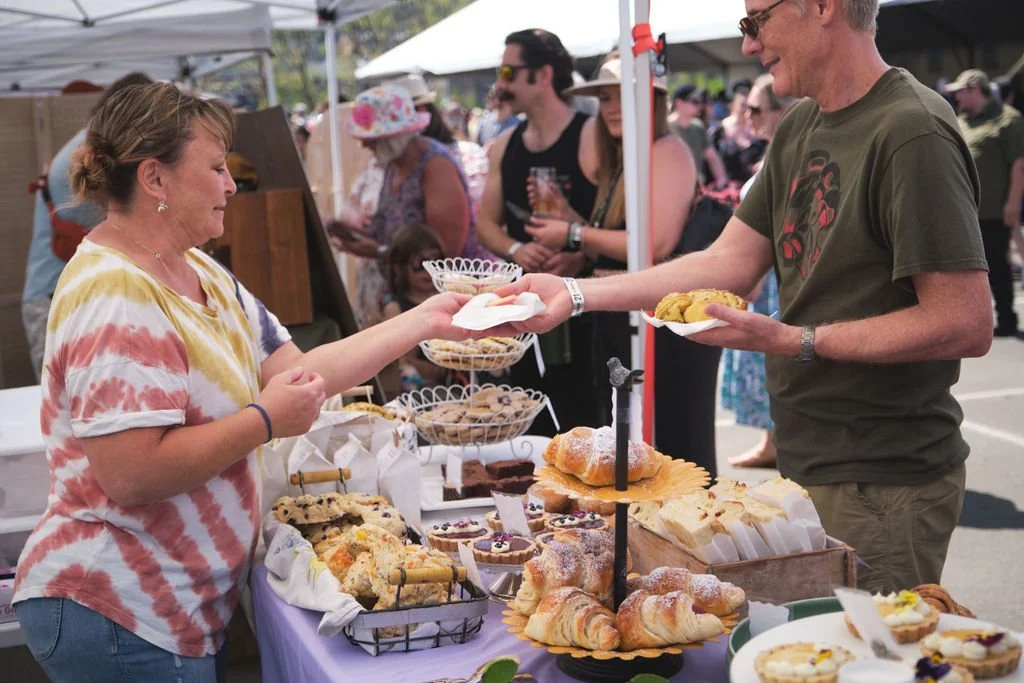 A woman at a bakery stall at an outdoor market handing a plate of baked goods to a man. The table has croissants, cookies, and pastries with a crowd of people in the background.
