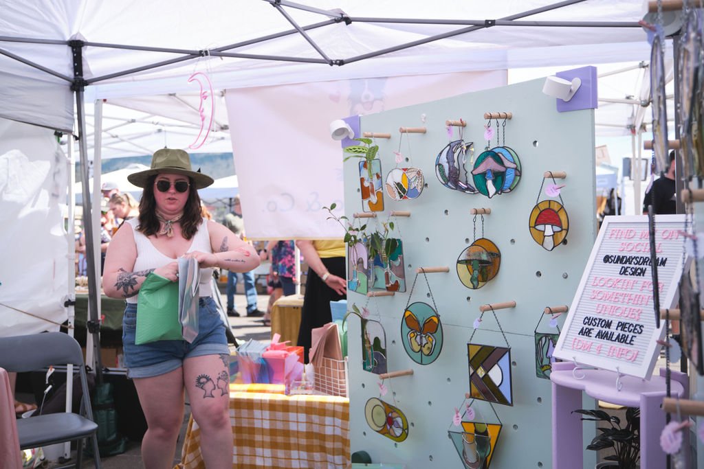 A woman with dark hair, sunglasses, a wide-brimmed hat, a tattoo on her arm and thigh, wearing a white sleeveless top and denim shorts, is browsing colorful handmade jewelry at an outdoor market stall.