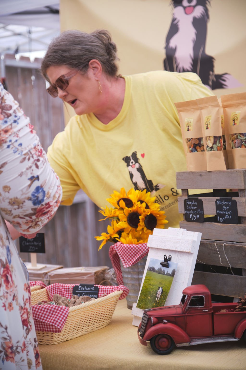 A woman at a market stall talking to a customer. She wears a yellow shirt with a dog graphic and sunglasses. The stall displays sunflower bouquets, bags of trail mix labeled pumpkin & lavender or blueberry oat, a wicker basket with items, a photo of 