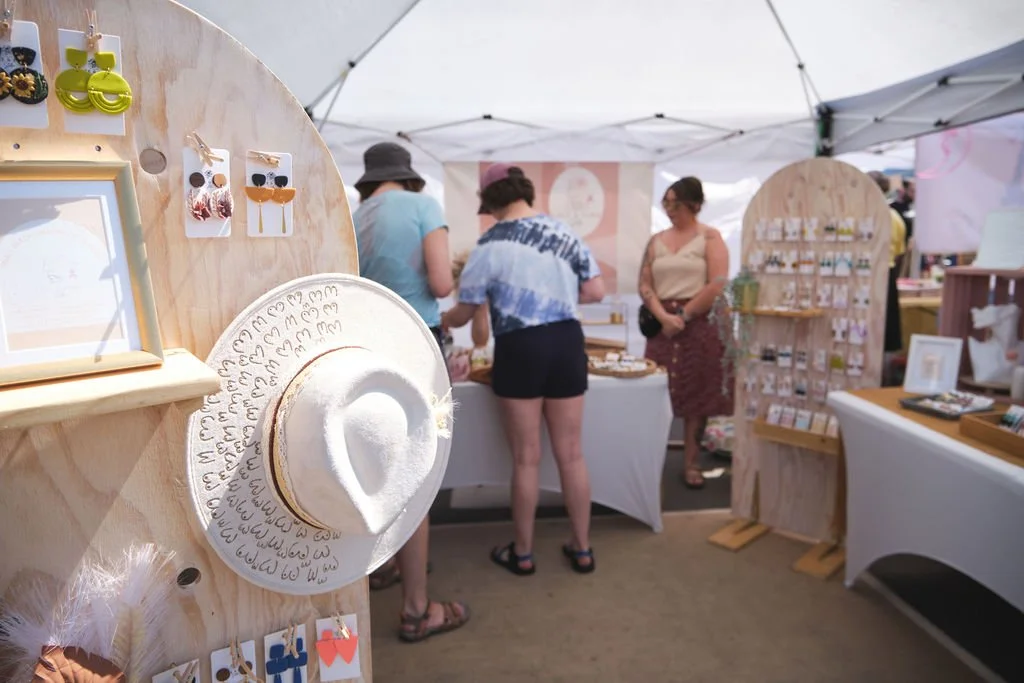 People shopping at a craft fair under a white tent, with jewelry, accessories, and decorative items displayed on tables and stands.