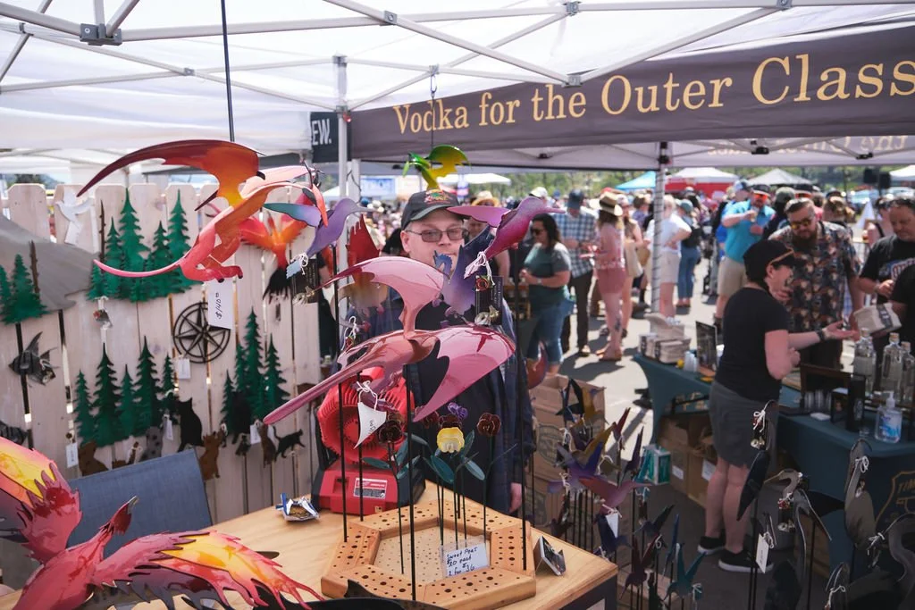 A man standing behind a booth at an outdoor market with colorful paper dragon decorations and a sign reading 'Vodka for the Outer Class.' There are other booths and shoppers in the background.