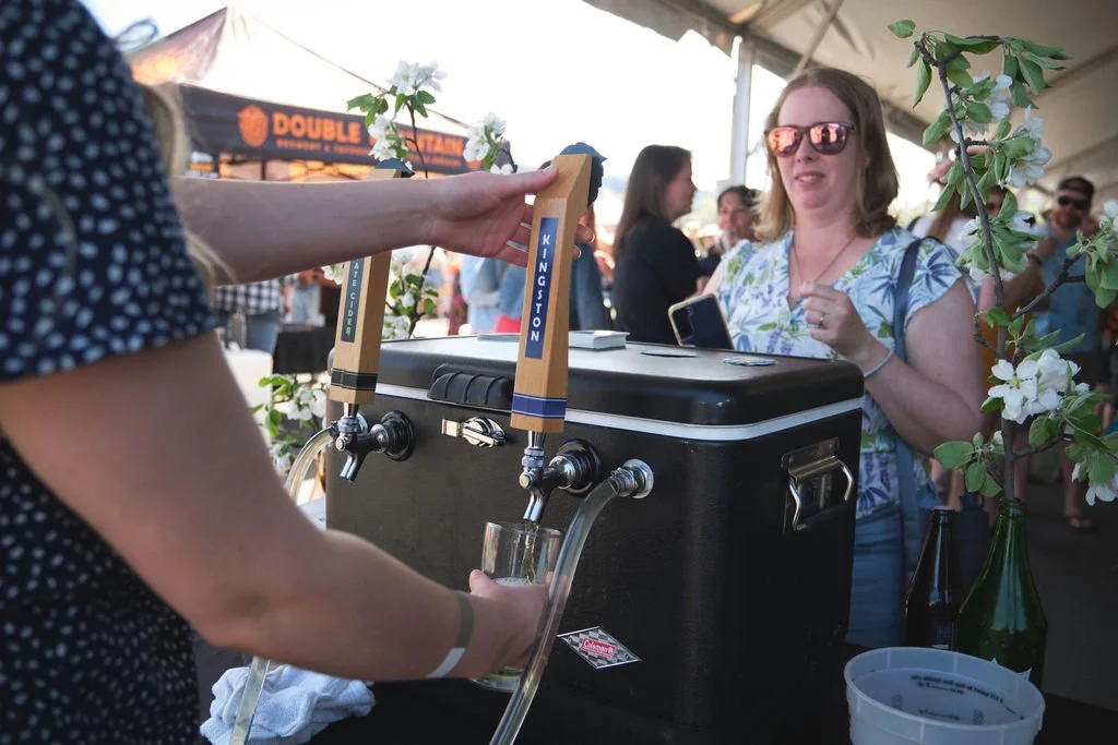 A woman in sunglasses and a floral shirt is at a bar, ordering from a tap system with two beer taps labeled 'Kingston' and 'Ate Green' at an outdoor event. People are in the background under a tent with a sign that reads 'Double.'