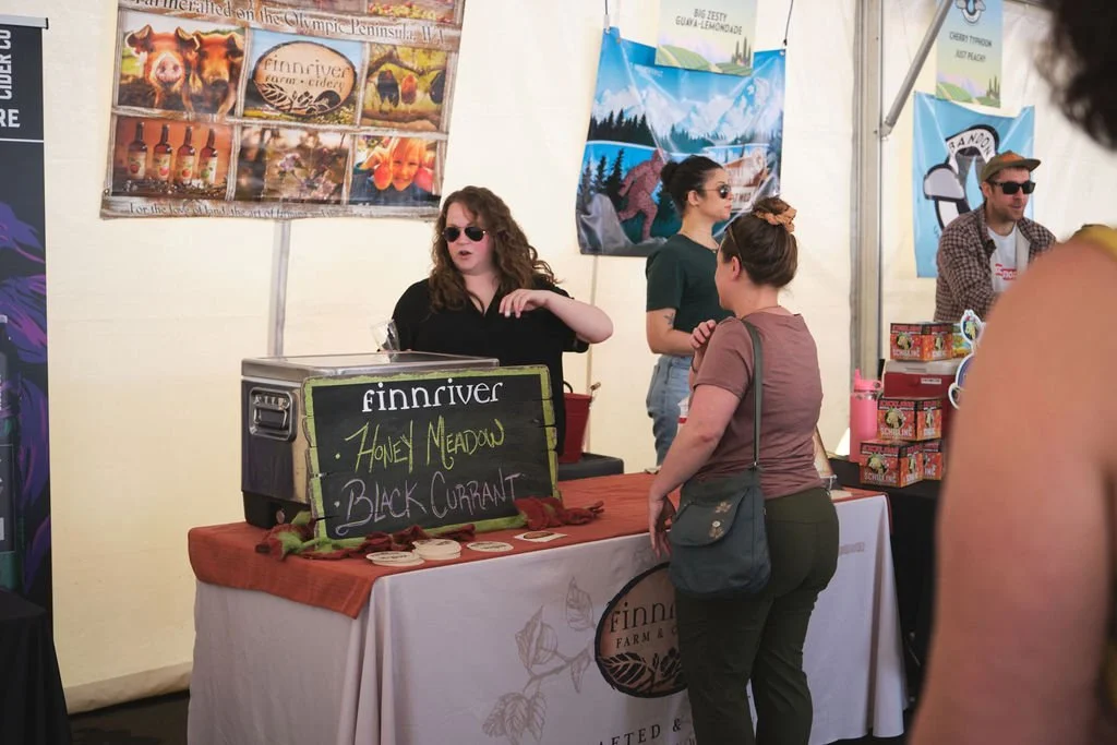A woman with curly hair and sunglasses talking to a vendor at a booth selling honey and blackberry jam at a fair or market.