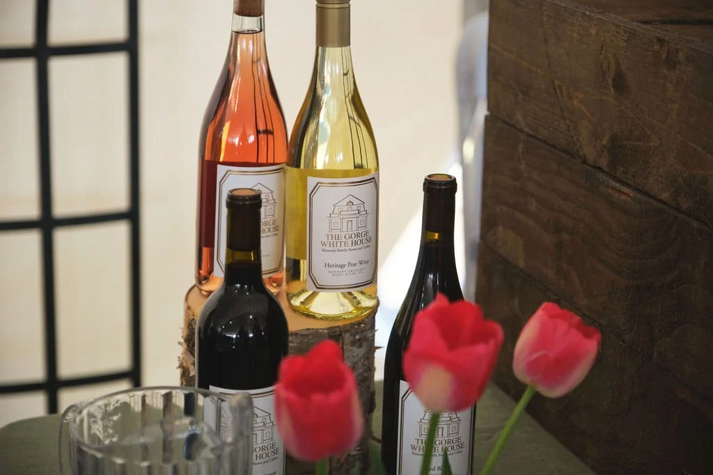 Four wine bottles, two with rosé, one with white wine, and one with red wine, positioned behind a glass and three pink tulips on a green table.