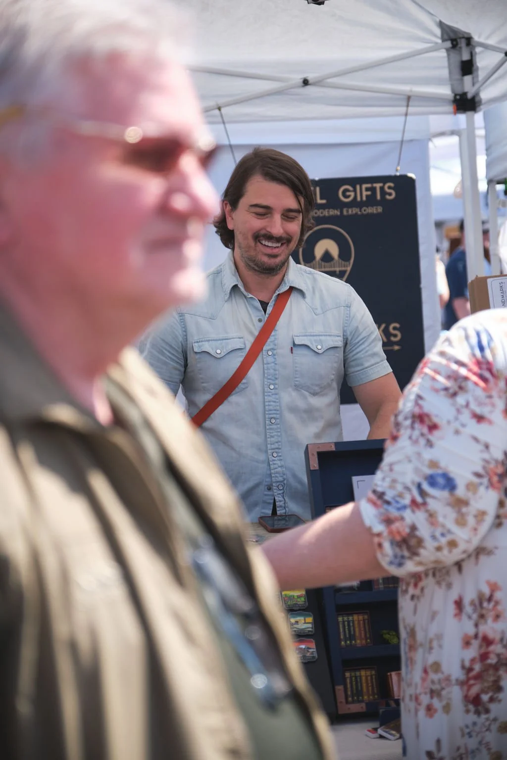A man with dark hair and a beard smiling at a market booth, wearing a light blue short-sleeve shirt with a brown strap across his chest. In the foreground, an out-of-focus older man with glasses and a striped shirt is visible, along with part of a wo