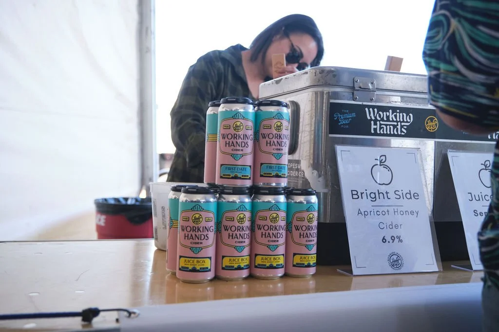 Cans of 'Working Hands' cider stacked on a table near a sign that reads 'Bright Side Apricot Honey Cider 6.9%', with a person in the background working at a food stand.