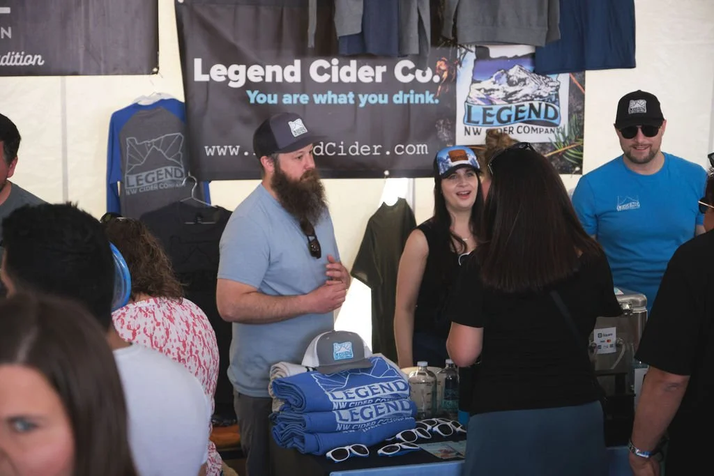 People chatting at a booth for Legend Cider Co. surrounded by merchandise like T-shirts, hats, and sunglasses, with a banner displaying the company's logo and slogan 'You are what you drink'.