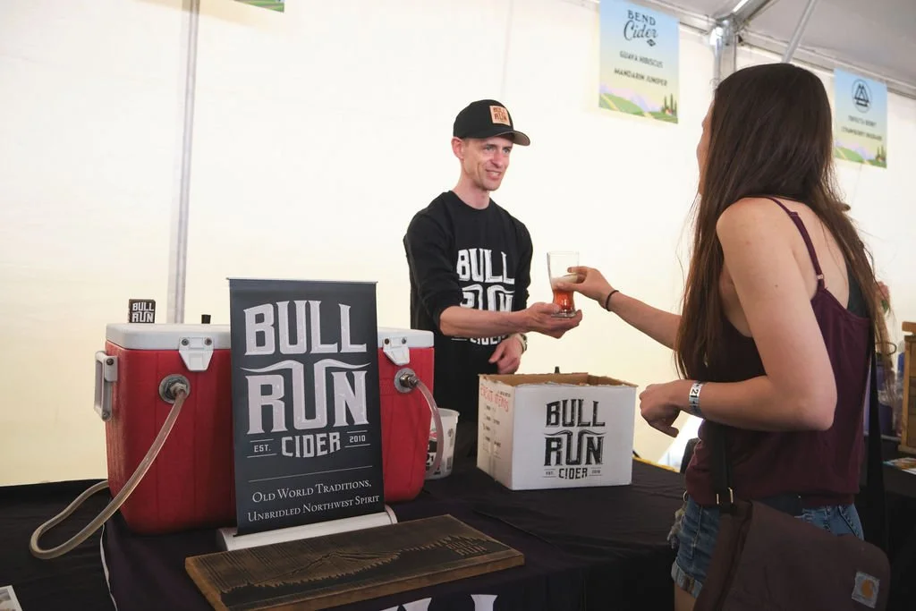 A woman in a maroon top and jeans receives a glass of cider from a man at a cider stand labeled 'Bull Run Cider.' The stand has a sign, a red cooler, and a cardboard box, with banners in the background.