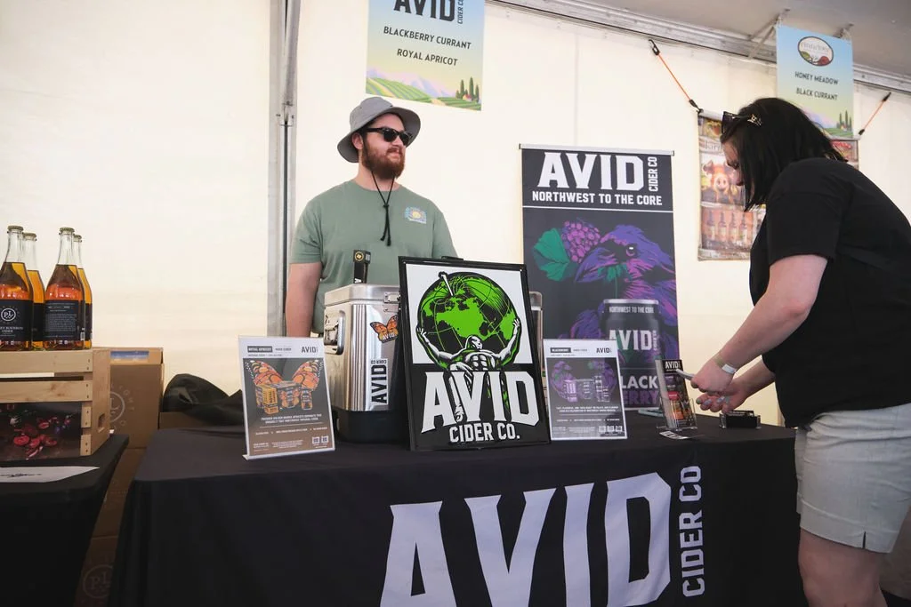 A man with a beard, sunglasses, and a sun hat stands behind a booth with the sign 'AVID Cider Co.' inside a tent. A woman in black shirt and beige shorts is at the booth, holding a phone. The booth displays promotional materials and bottles of cider,