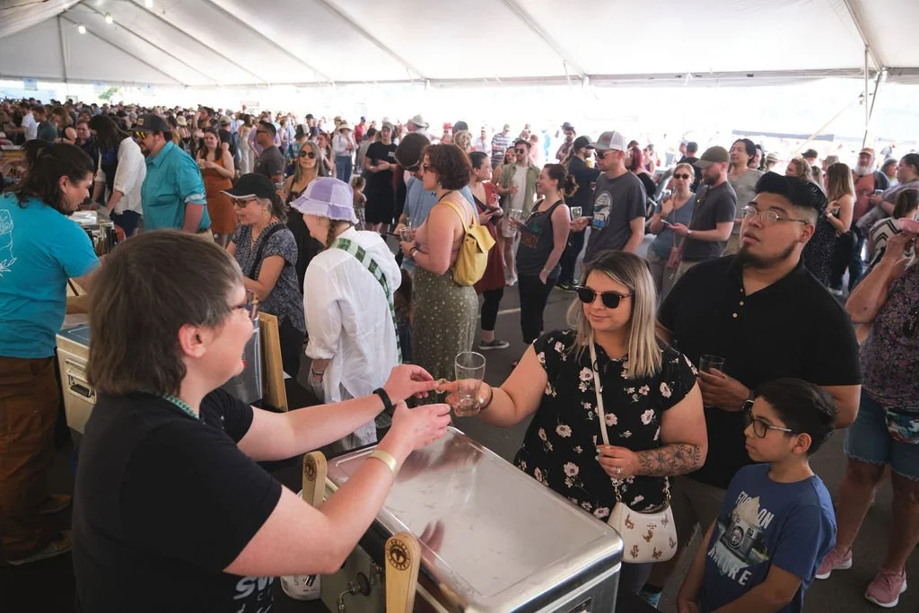 People attending a crowded outdoor event under a large white tent, standing in line and socializing.
