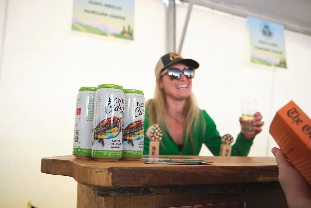 A woman in a green jacket, sunglasses, and a cap is smiling and holding a drink behind a wooden booth. Several cans of Bend Cider with colorful labels are on the counter in front of her.