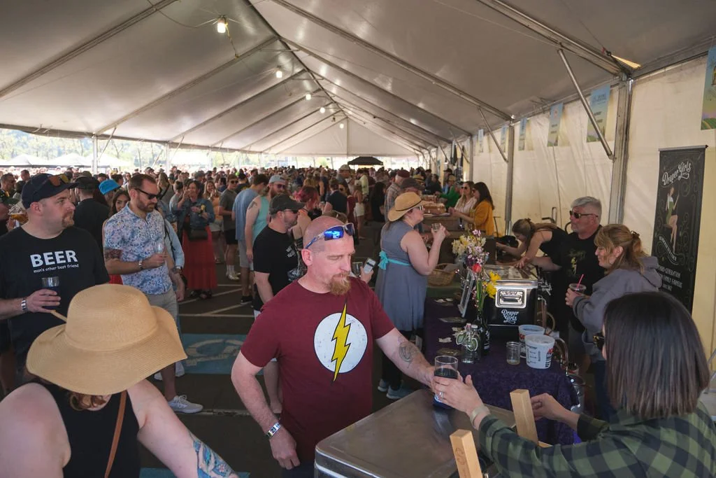 Crowd of people gathered inside a large event tent, some standing in line at a food or drink stand with servers, colorful decorations, and string lights hanging from the ceiling.