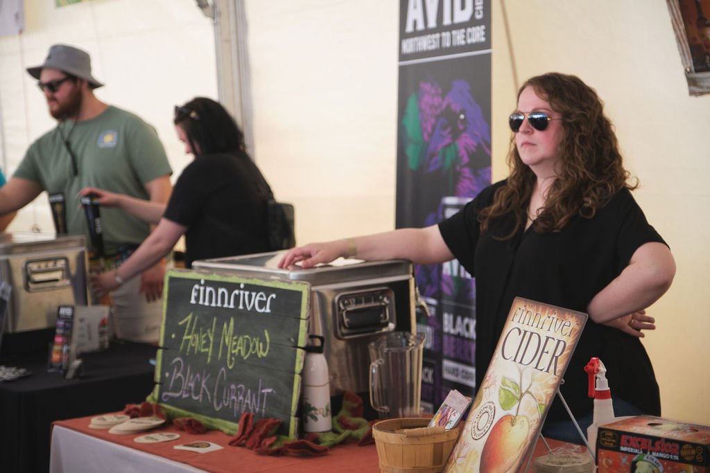 A woman with curly brown hair wearing sunglasses and a black shirt standing behind a table selling cider at a festival or market. The table has a sign with the words "finniiver" and mentions "Ashley Meadow" and "Black Currant." A poster behind her di