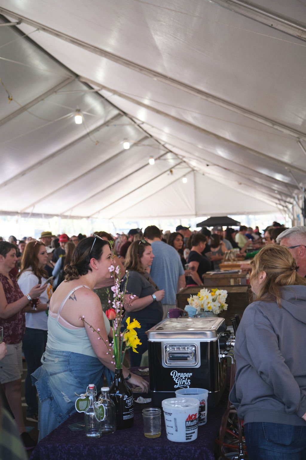 Crowd of people inside a large event tent, with some standing near or at a food or drink stall decorated with flowers and beverages.