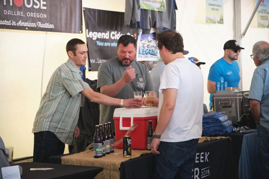 Three men standing behind a table with bottled drinks and a red cooler, engaging in conversation at a beer event or festival.