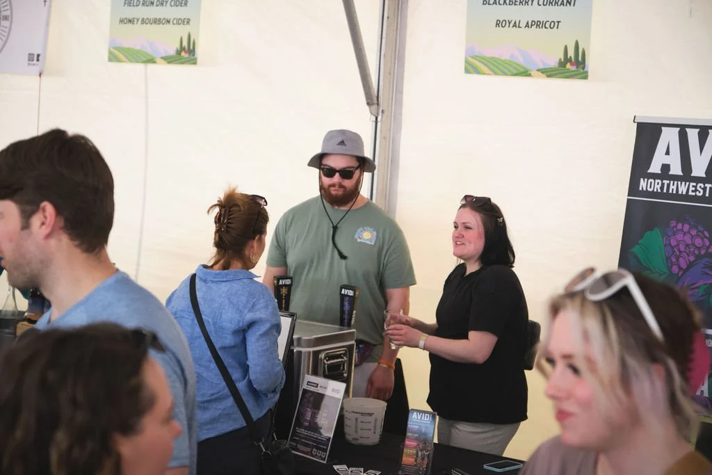 A group of people at a booth or stand in an indoor event or festival, with posters and banners advertising cider flavors like honey bourbon cider, field run dry cider, blackberry currant, and royal apricot.