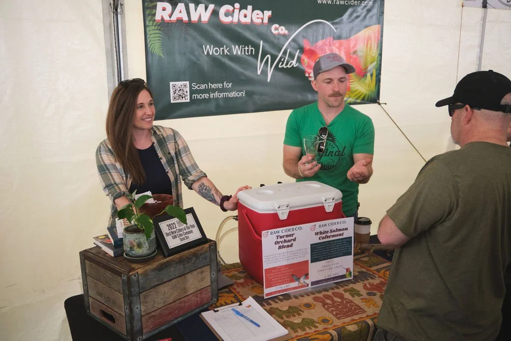 Two people are at a booth selling raw cider. The woman on the left is smiling and has long brown hair, and the man on the right has a mustache and is wearing a green t-shirt and a gray cap. They are engaging with a customer. Behind them is a banner t