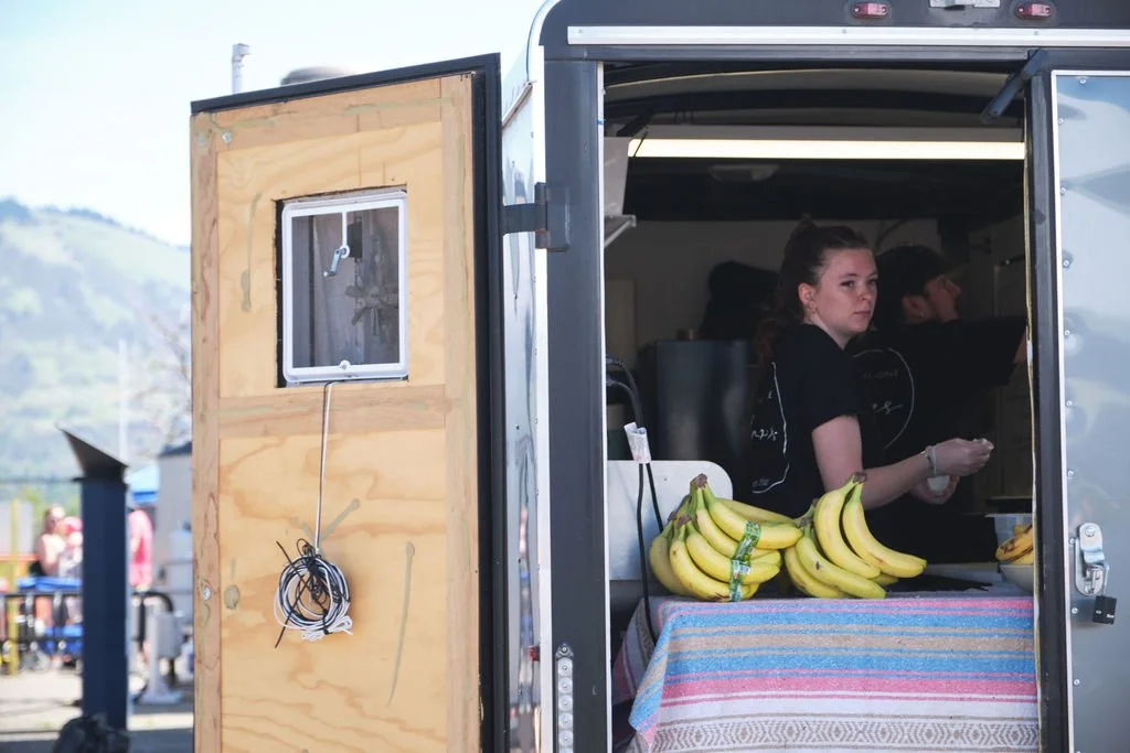 A food truck serving bananas with a young woman working inside, and people in the background.