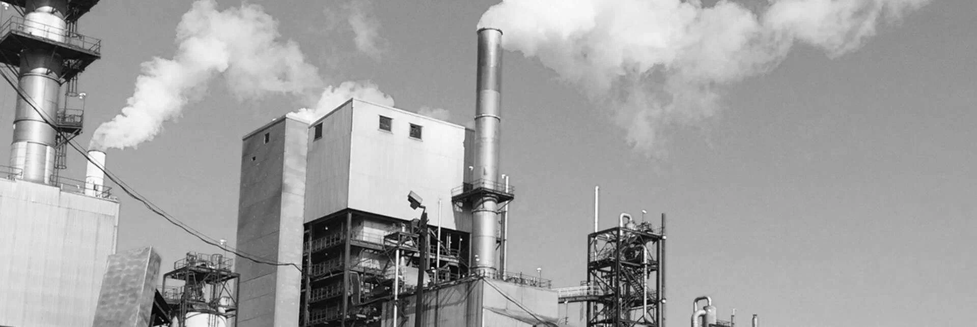 Industrial factory with multiple smokestacks emitting white smoke, metal structures, and pipes against a partly cloudy sky.